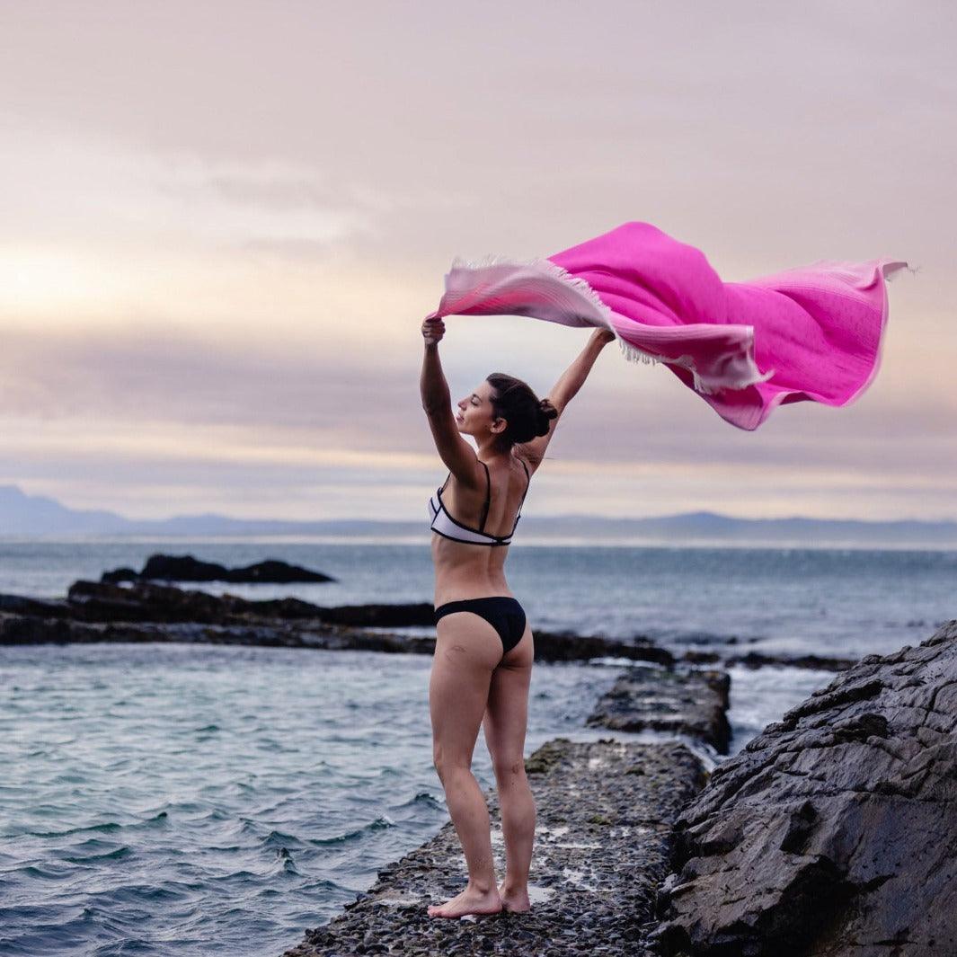 Lady holds overhead vivid pink towel by the seashore