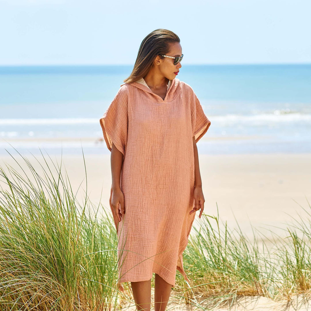 A woman at the beach wearing a coral colour beach changing robe, with hood down.